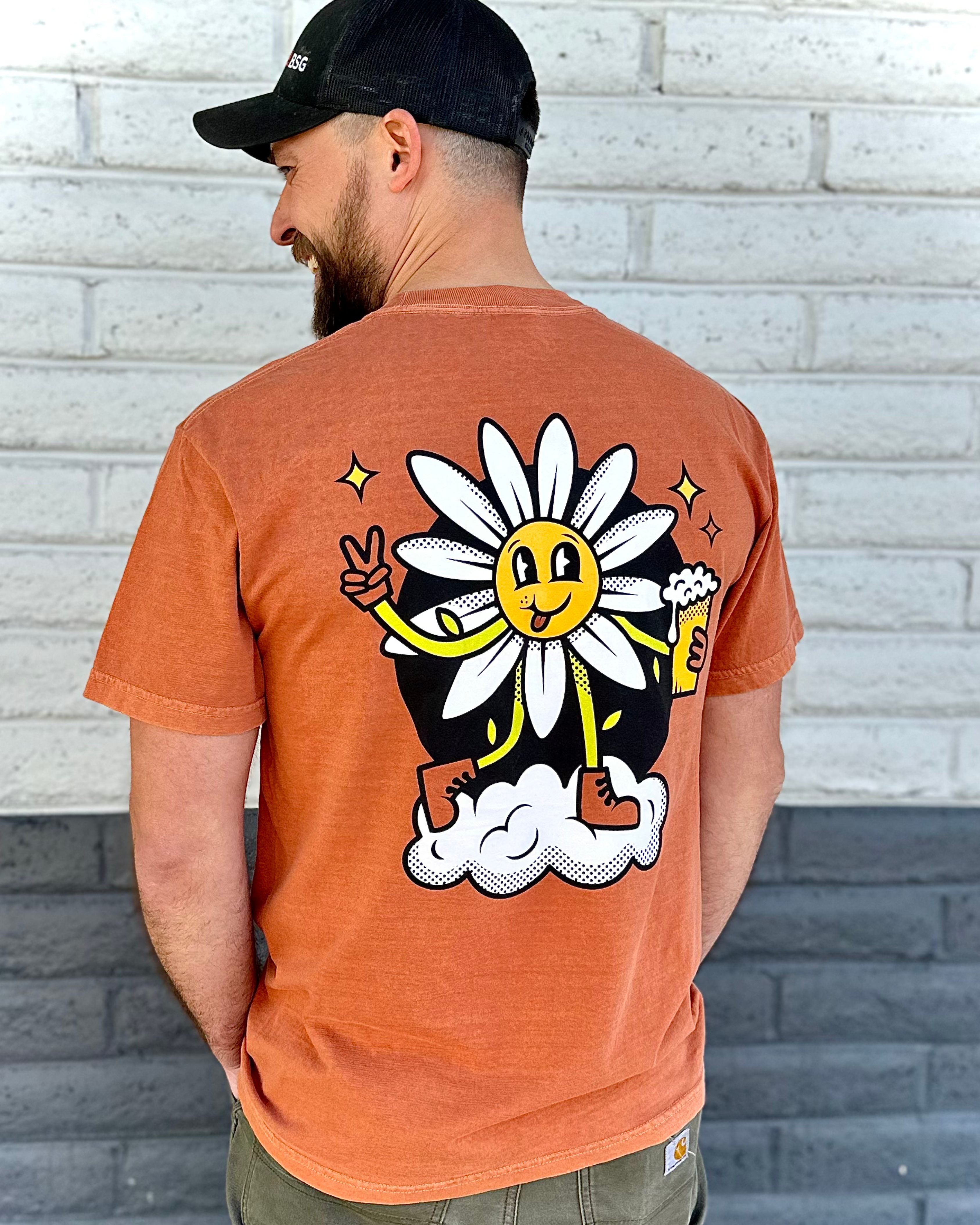 Man wearing an orange t-shirt with a cartoon daisy design against a brick wall.